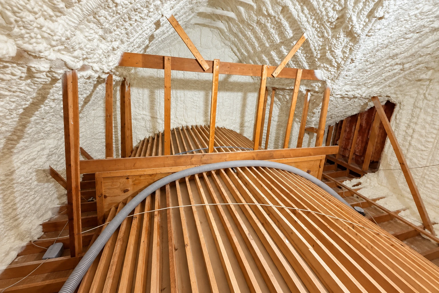 Attic framing and rafters are surrounded by thick white spray foam insulation, with a flexible hose across the joists.