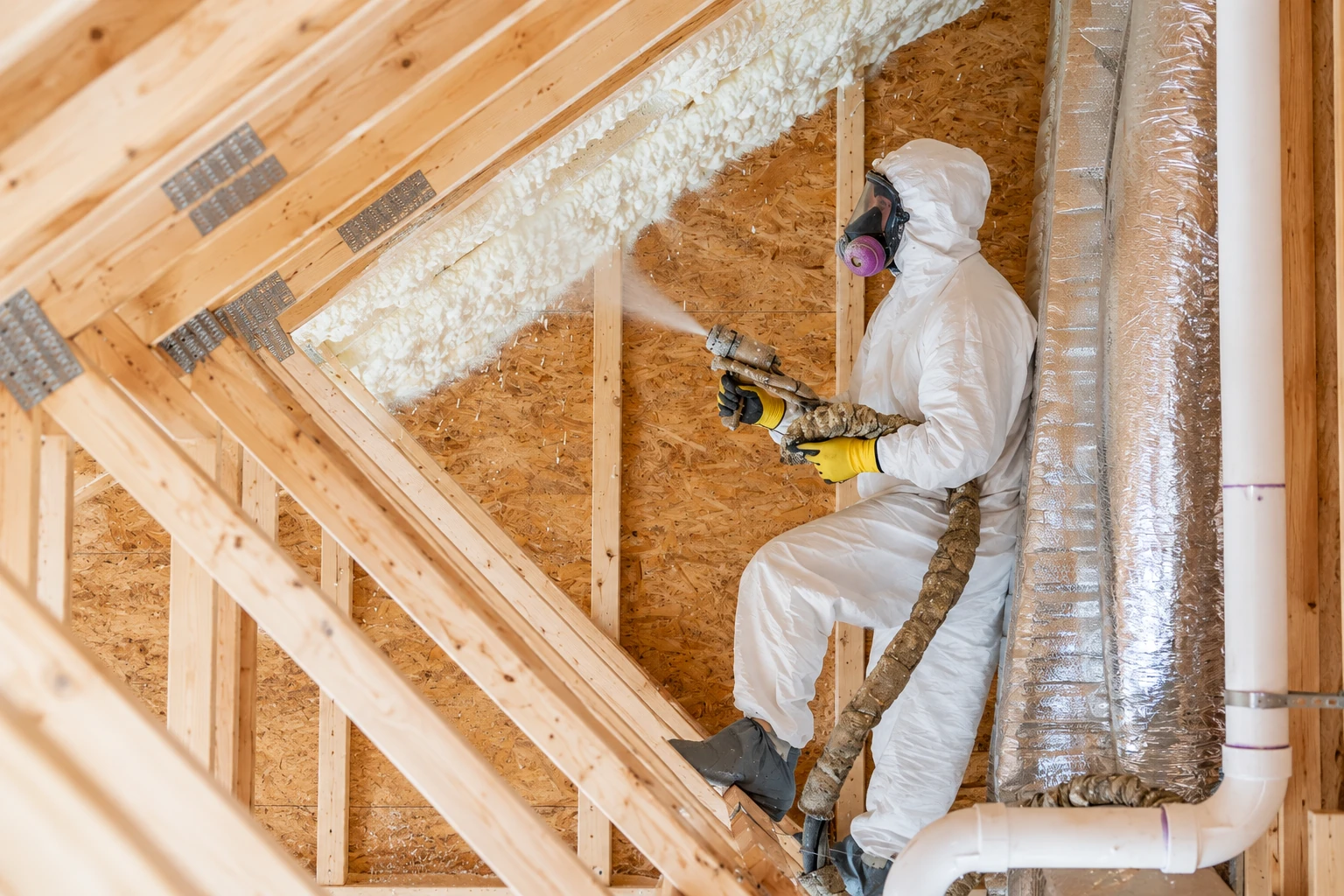 A worker in protective gear sprays foam insulation between the exposed wooden framing of an attic.