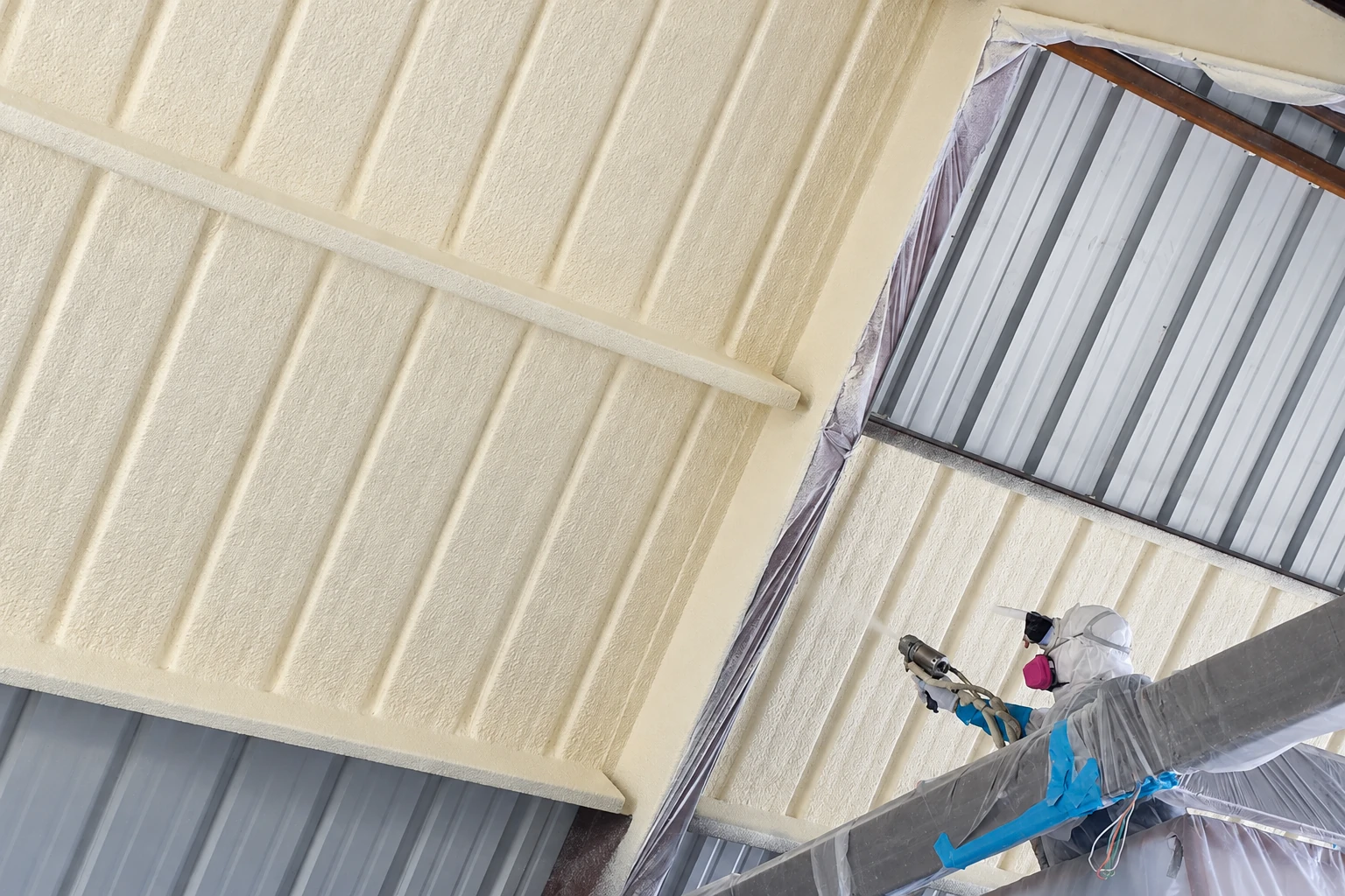 A worker in protective gear sprays pale closed-cell foam onto the underside of a corrugated metal roof.