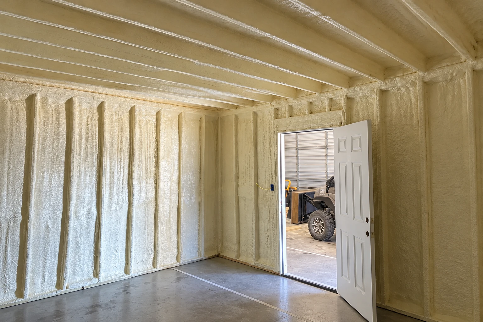 A commercial room has pale spray foam covering its walls and ceiling, with an open door to a garage.