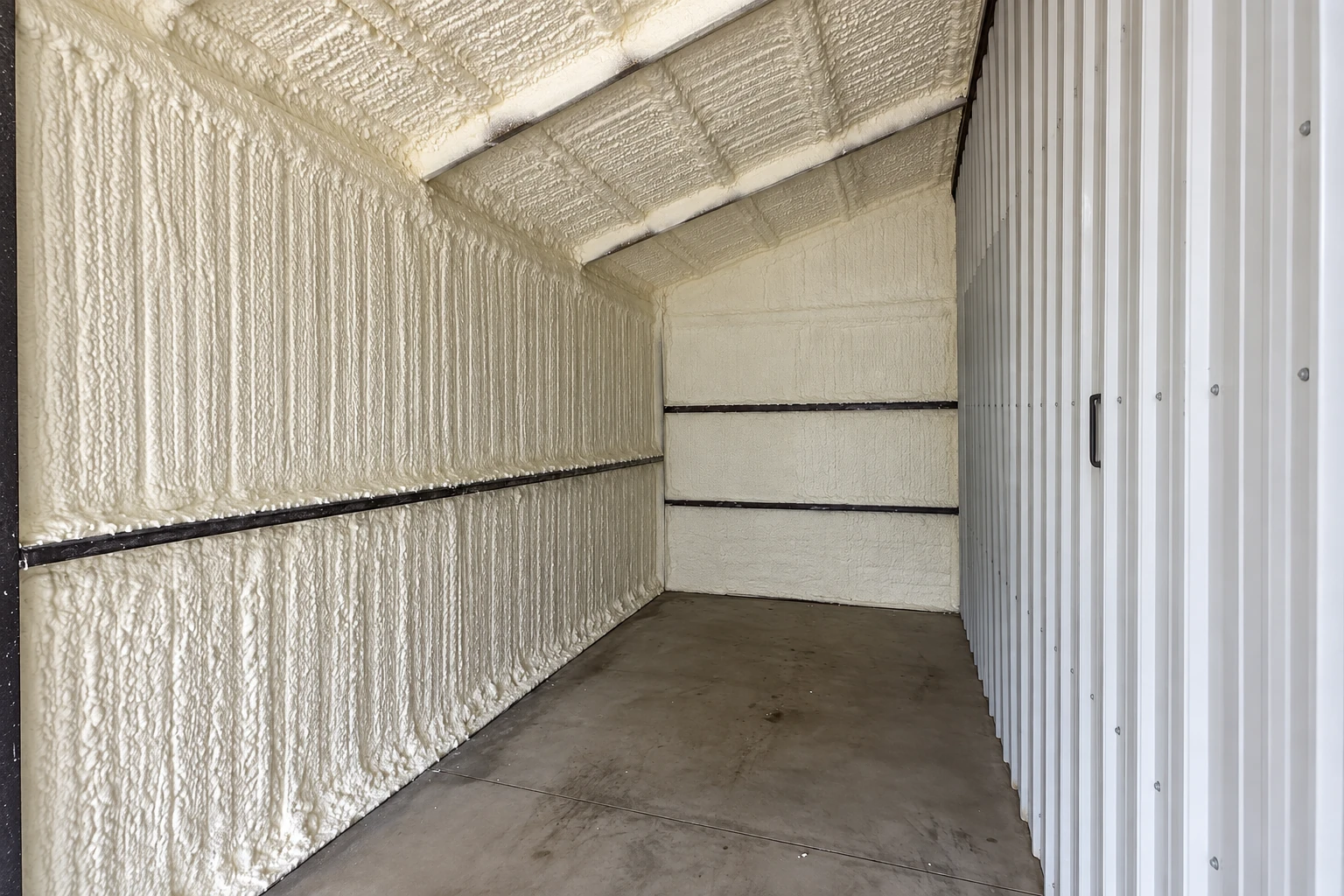 A narrow commercial space has white spray foam covering its walls and sloped ceiling, with a concrete floor and metal paneling.