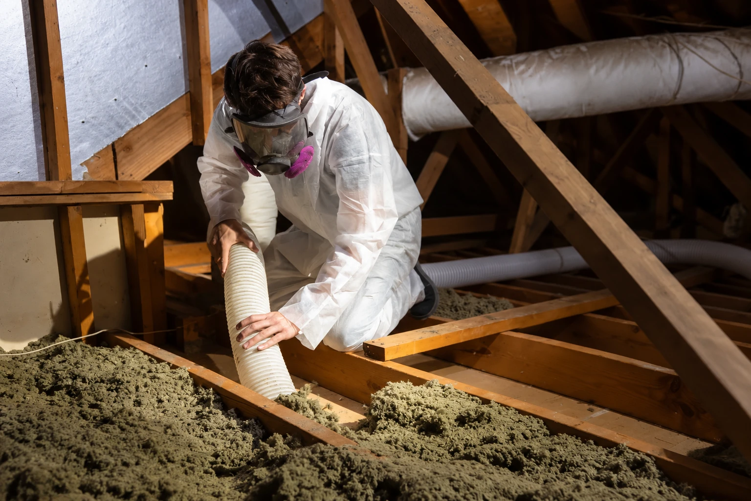 A worker in protective coveralls and a respirator removes attic insulation using a flexible vacuum hose.