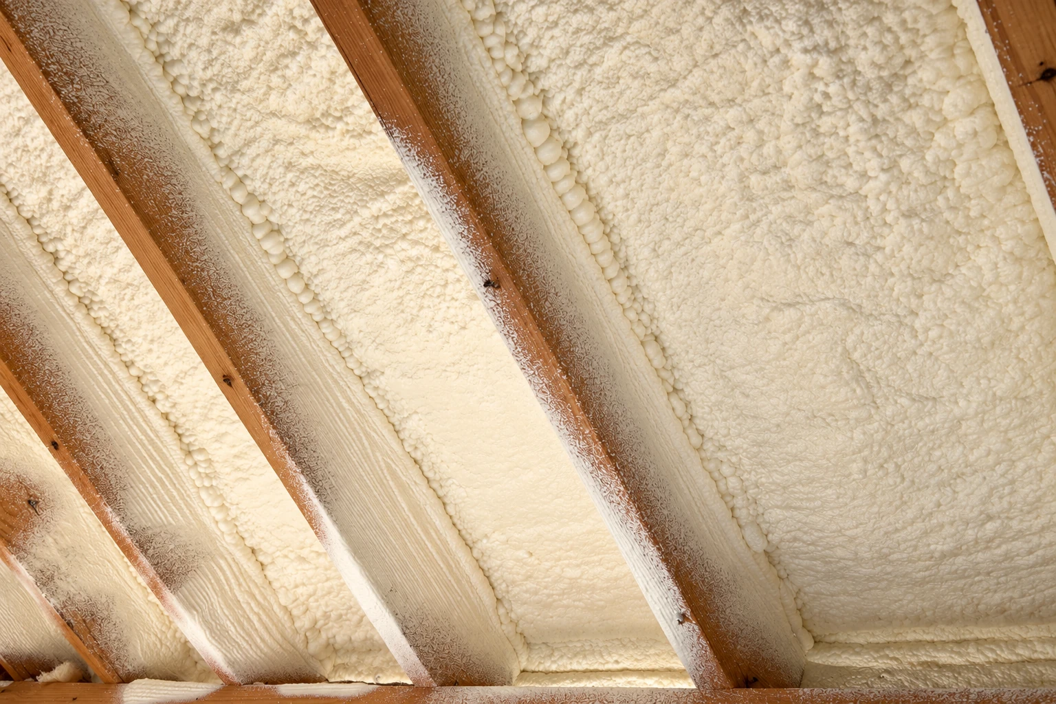 Cream-colored spray foam fills the spaces between exposed wooden rafters on a ceiling.