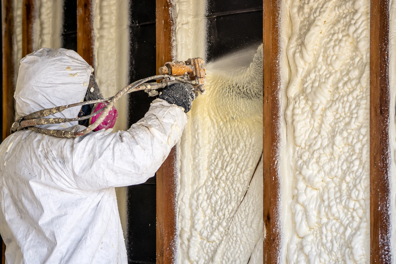 Worker in protective coveralls sprays foam insulation into wall cavities between exposed wooden studs.